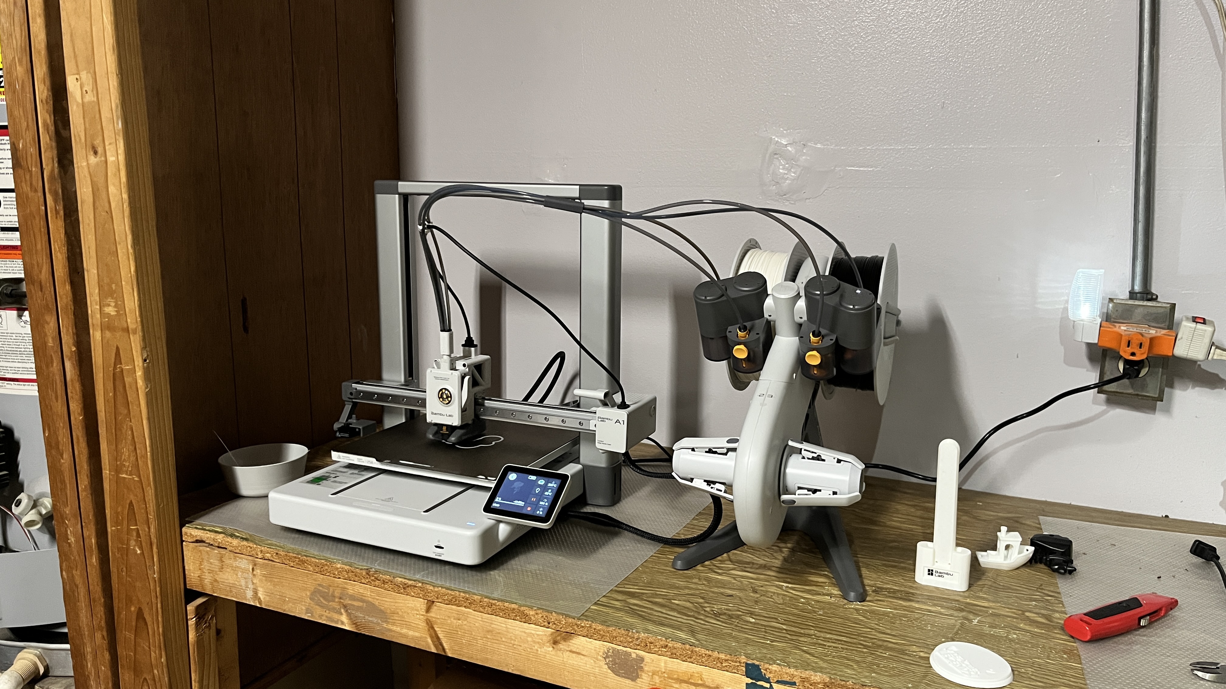 A Bambu Lab A1 3D printer setup on a wooden workbench, with a filament AMS (Automatic Material System) unit connected via tubes for multi-material printing. The printer's touchscreen interface is visible, showing the status. Several 3D printed objects, including a Benchy boat, calibration cube, and other prints, are scattered on the right side of the bench near a red utility knife. The setup is located in a workshop with wooden paneling and a power outlet with a nightlight and orange extension plug.